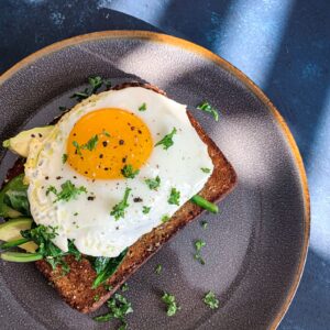 A plate with avocado toast topped with a sunny-side-up egg and sprinkled with chopped parsley. The plate has a dark rim and rests on a blue surface with shadows.