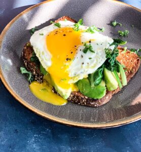 A plate with avocado toast topped with sautéed greens and a sunny-side-up egg, with yolk oozing over the sides. The dish is garnished with fresh herbs and served on a speckled ceramic plate against a dark surface.