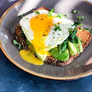 A plate with avocado toast topped with sautéed greens and a sunny-side-up egg, with yolk oozing over the sides. The dish is garnished with fresh herbs and served on a speckled ceramic plate against a dark surface.