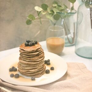 A stack of pancakes topped with blueberries and drizzled with syrup on a white plate. More blueberries are scattered around. In the background, theres a glass of coffee and a green plant in a vase. A beige napkin and a fork are in the foreground.