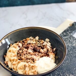 A bowl of oatmeal topped with sliced bananas, nuts, and a dollop of yogurt, arranged on a gray stone platter with a marble background.