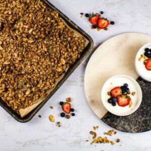 A baking tray with granola on parchment paper is on the left. On the right, two bowls of yogurt topped with strawberries, blueberries, and granola sit on a round cutting board. Scattered fruit and granola bits decorate the marble surface.