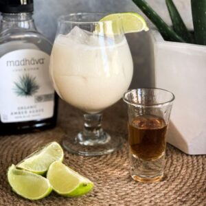 A glass filled with a creamy beverage garnished with a lime wedge sits on a woven mat. Nearby are sliced limes, a shot glass containing a dark liquid, a bottle labeled Madhava Organic Agave, and an aloe plant in a white pot.