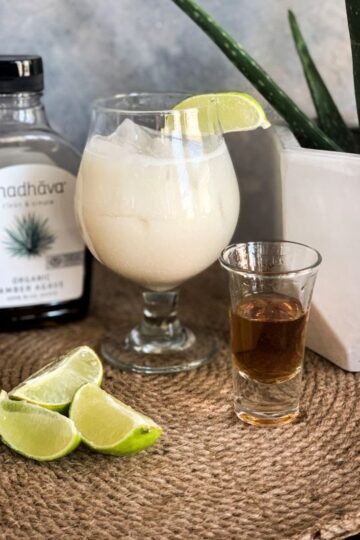 A glass filled with a creamy beverage garnished with a lime wedge sits on a woven mat. Nearby are sliced limes, a shot glass containing a dark liquid, a bottle labeled Madhava Organic Agave, and an aloe plant in a white pot.