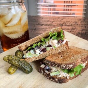 A wooden board displays two chicken salad sandwiches with lettuce and cranberries on whole grain bread, following a delightful cranberry chicken salad recipe. Three small pickles rest beside them, while in the background, a mason jar brims with refreshing iced tea.