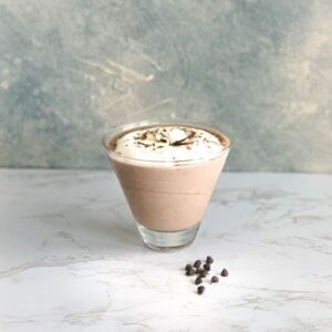 A glass of chocolate mousse topped with whipped cream and chocolate shavings sits on a marble surface. A few chocolate chips are scattered nearby. The background is a textured grey and white wall.