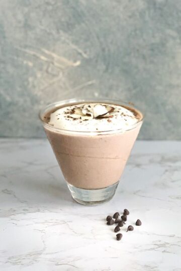 A glass of chocolate mousse topped with whipped cream and chocolate shavings sits on a marble surface. A few chocolate chips are scattered nearby. The background is a textured grey and white wall.