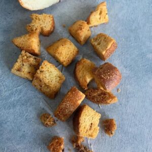 Scattered croutons on a textured gray surface, with a partially visible loaf of bread in the top corner. The croutons are golden brown and vary in size.