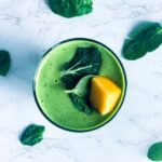 A green smoothie in a glass, garnished with fresh spinach leaves and a mango chunk, sits on a marble surface. Spinach leaves are scattered around the glass.