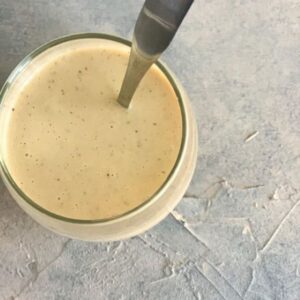 A glass filled with creamy smoothie, light beige in color, sits on a textured gray surface. A metal spoon is inserted into the glass. Small specks are visible within the smoothie, suggesting ingredients like vanilla or spices.