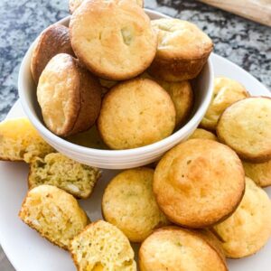 A white plate is filled with a variety of golden-brown muffins, some displayed in a bowl. A few muffins are cut open, revealing a soft, moist interior with small bits of green and yellow vegetables. The background shows a granite countertop.