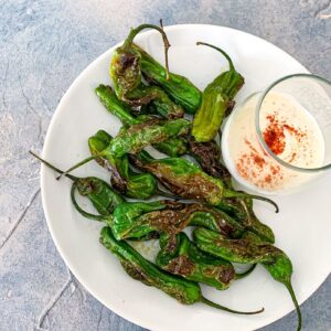 A white plate of charred shishito peppers with a small glass bowl of creamy dip sprinkled with paprika, on a textured gray background.