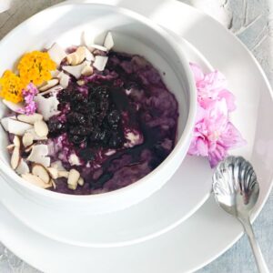 A white bowl of purple porridge topped with dried fruit, sliced almonds, coconut flakes, and yellow edible flowers. Its placed on a white plate with pink flowers on the side and a silver spoon next to it.