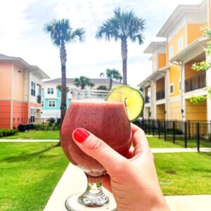 A hand with red nail polish holds a glass of smoothie garnished with a lime slice. The background features colorful apartment buildings, green grass, and palm trees under a partly cloudy sky.