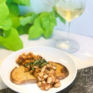 A plate with toasted bread topped with a bean and herb mixture. In the background, a glass of white wine and green leafy plants are visible, creating a fresh and elegant dining scene.