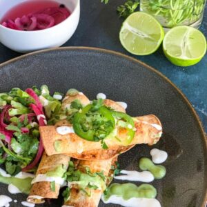 A plate of rolled tacos topped with jalapeño slices, drizzled with creamy sauce, and served with a side of salad. Pickled onions in a bowl and lime halves accompany the dish. Fresh cilantro is visible in the background.