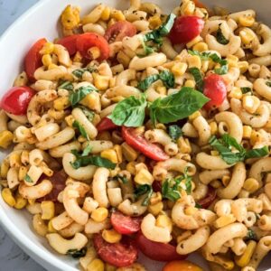 A bowl of pasta salad with elbow macaroni, cherry tomatoes, corn, and basil leaves. The dish is seasoned with spices and herbs, creating a colorful and fresh appearance on a light marble surface.