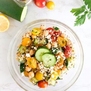 A fresh salad in a glass bowl featuring cherry tomatoes, chickpeas, sliced cucumber, quinoa, and crumbled feta cheese. A lemon, cucumber, and parsley are nearby on a white marble surface.