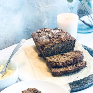 A sliced loaf of homemade banana bread on a wooden board, accompanied by a bowl of butter. A lit candle and a vase with green leaves are in the background, creating a cozy atmosphere.