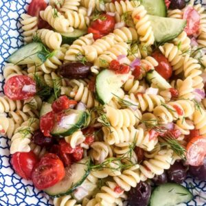 A vibrant pasta salad featuring rotini, cherry tomatoes, cucumbers, black olives, red onions, and fresh dill, all tossed together in a blue and white patterned bowl.