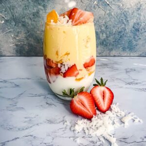 A layered dessert in a glass features white yogurt, red strawberries, orange mango puree, and shredded coconut. Additional strawberry slices and coconut are scattered in front of the glass on a textured surface.