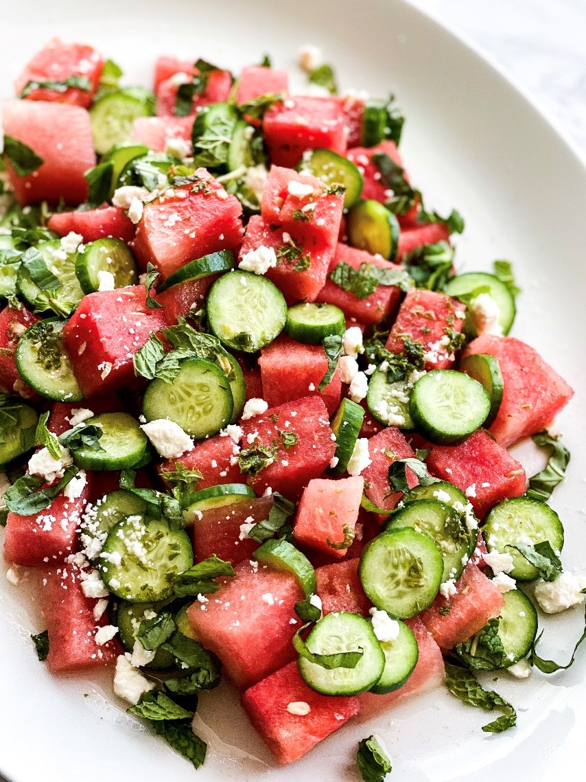 A fresh salad featuring cubed watermelon, sliced cucumbers, and crumbled feta cheese, garnished with chopped mint leaves, arranged on a white plate.