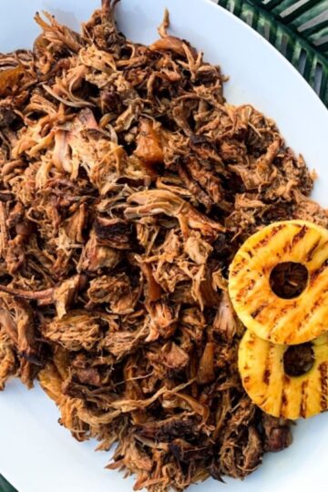 Pulled pork on a white platter accompanied by grilled pineapple slices, placed on a green woven surface.