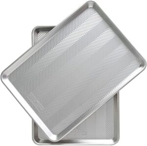 Two metal baking sheets with a chevron pattern on their surfaces are stacked slightly offset from each other. They have raised edges and are likely used for baking or roasting.