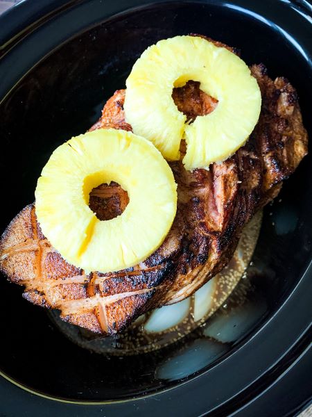 A glazed pork roast garnished with two pineapple rings, placed in a black roasting dish. The ham is scored and cooked to a golden brown.