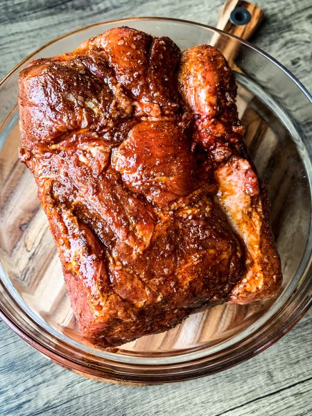 A marinated piece of pork sits in a glass bowl on a wooden cutting board. The meat has a rich, reddish-brown color with visible seasonings, suggesting it is well-seasoned and ready for cooking.