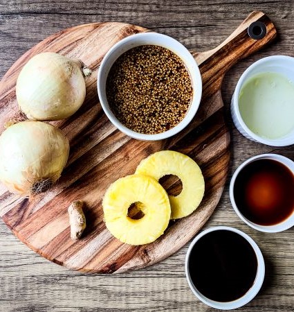 Ingredients for Hawaiian Pulled Pork braising liquid on a wooden board: two onions, three pineapple slices, a piece of ginger, a bowl of mustard seeds, and three small bowls containing green sauce, dark brown liquid, and light brown liquid.