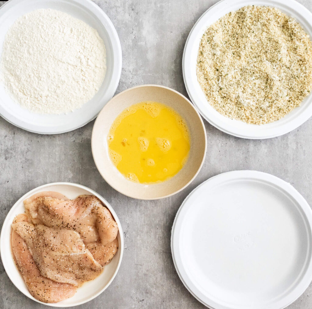 a dredging station with 5 plates with chicken, flour, breadcrumbs, eggs, and an empty plate 