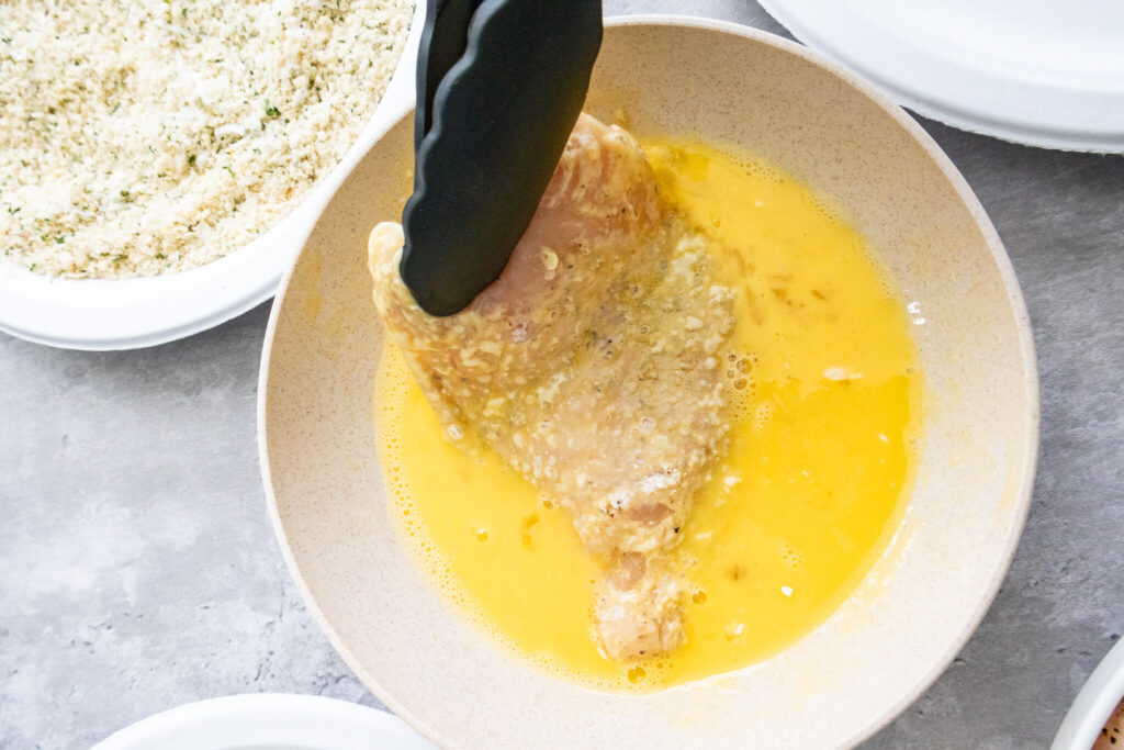 a pair of tongs dipping a chicken breast into whisked eggs  