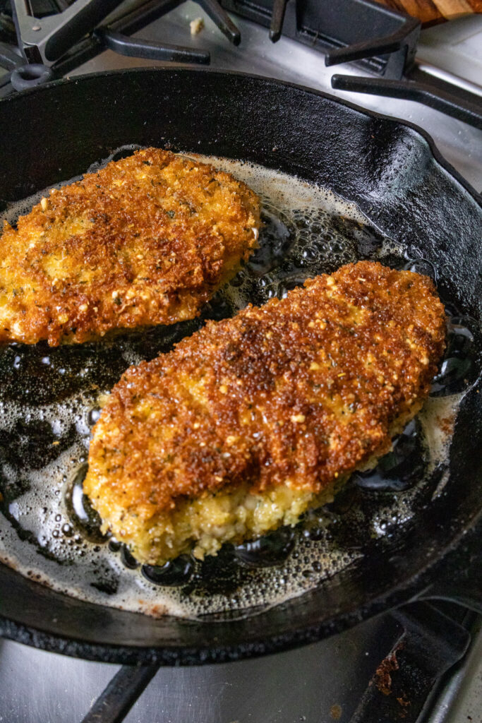 2 crispy chicken cutlets in a cast iron pan frying in oil