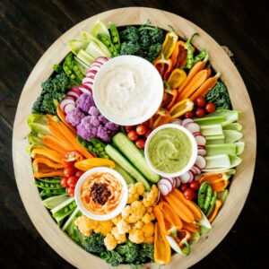 A colorful vegetable tray featuring a variety of fresh vegetables, including carrots, celery, cherry tomatoes, and bell pepper strips, arranged around three bowls of dips. The dips include a Classic Vegetable Dip, Caramelized Onion and Harissa Dip, and an Avocado Hummus.