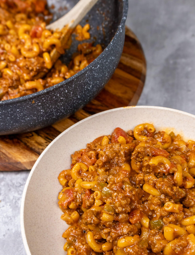 A serving of One-Pot Classic American Goulash in a bowl with the pan in the background. 