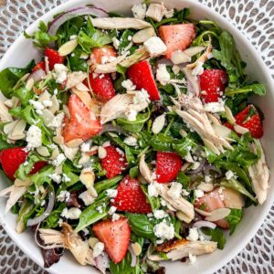 A vibrant strawberry poppy seed salad featuring mixed greens, spinach, sliced strawberries, shredded chicken, feta cheese, almond slices, and red onion on a white patterned plate. Perfect with a drizzle of poppy seed dressing.