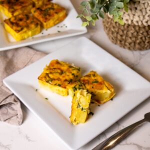 Three savory quiche pieces with vegetables and herbs are elegantly served on a white square plate. A beige cloth napkin and a woven pot plant add charm in the background, evoking a Starbucks vibe reminiscent of their copycat recipe for potato and chive egg squares. A fork rests nearby on marble.