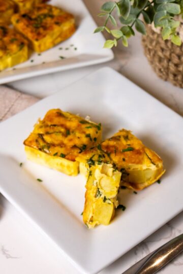 Three savory quiche pieces with vegetables and herbs are elegantly served on a white square plate. A beige cloth napkin and a woven pot plant add charm in the background, evoking a Starbucks vibe reminiscent of their copycat recipe for potato and chive egg squares. A fork rests nearby on marble.