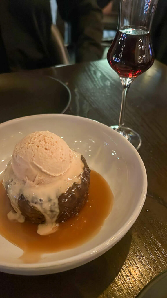 A dessert with a scoop of ice cream atop a sticky toffee pudding is served in a white bowl, surrounded by a caramel sauce. A glass of red wine is in the background on a dark wooden table.