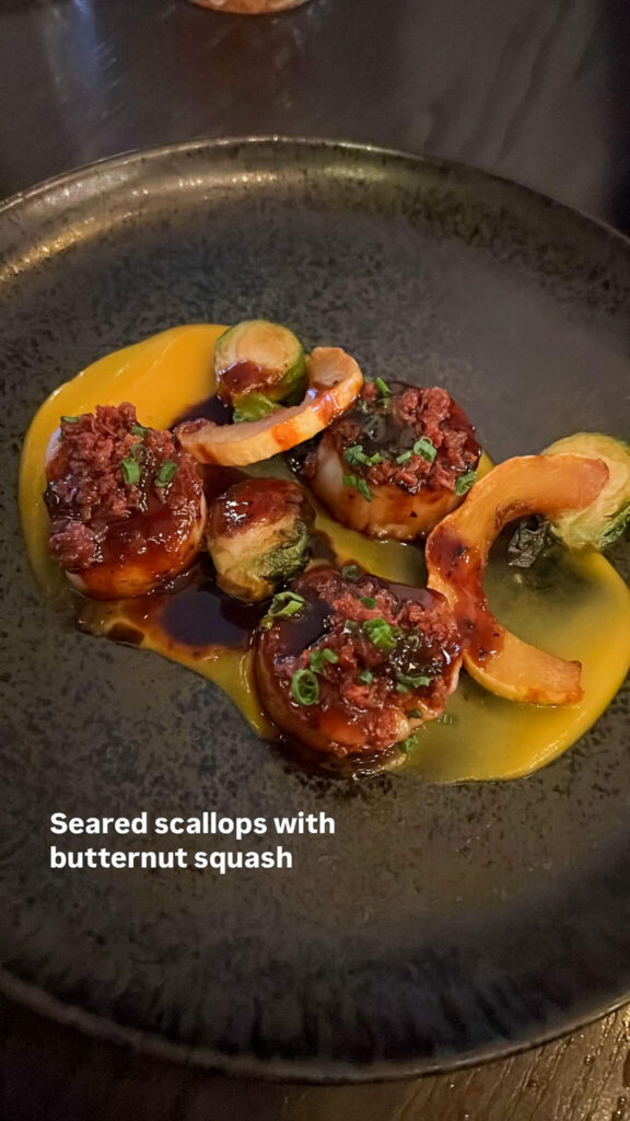 A dark plate with seared scallops topped with a savory sauce, garnished with herbs. Accompanied by roasted butternut squash slices and Brussels sprout halves on a swirl of butternut puree. Text on the image reads, Seared scallops with butternut squash.