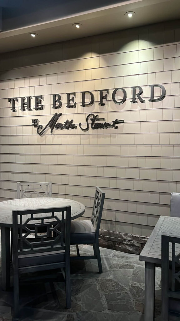 Restaurant interior featuring a sign reading The Bedford by Martha Stewart on a tiled wall. The foreground shows a round wooden table with cushioned chairs on a stone floor, under soft overhead lighting.