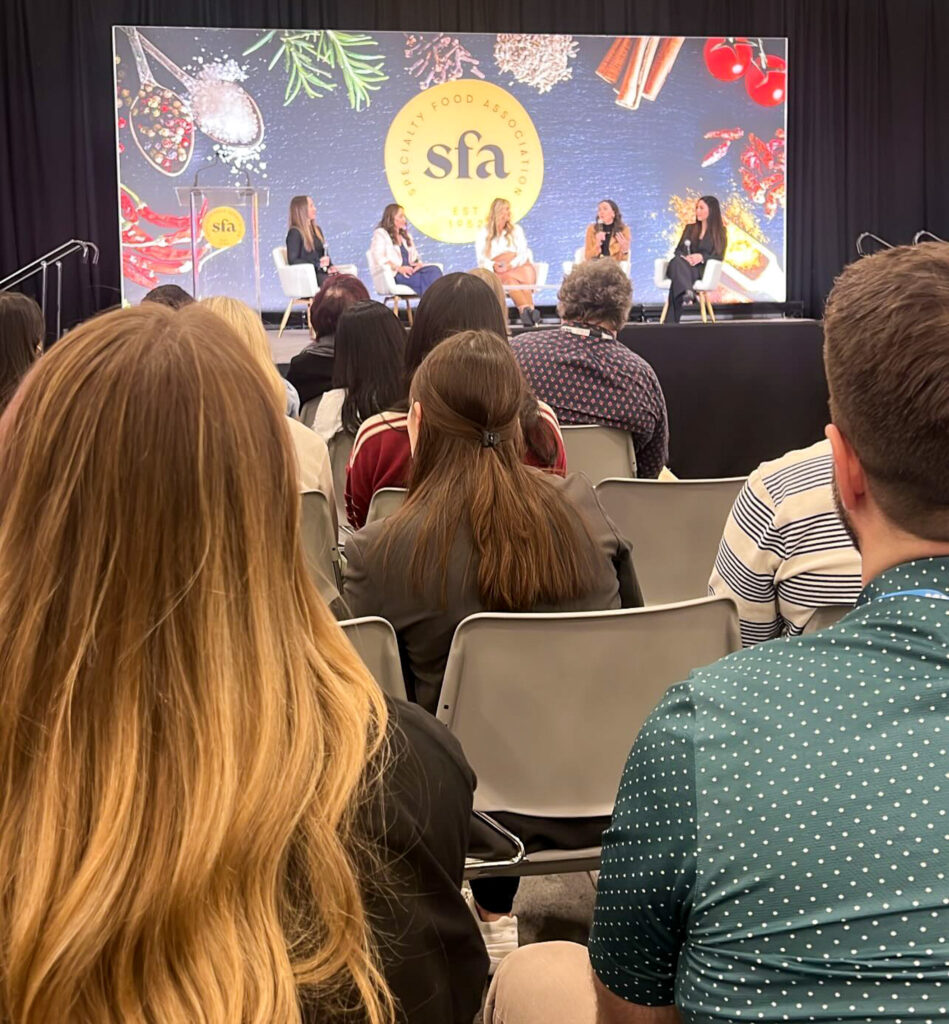 Audience members seated at a panel discussion event with five people on stage. The backdrop displays sfa and images of spices and herbs. The scene captures a diverse group of attendees and panelists engaged in conversation.