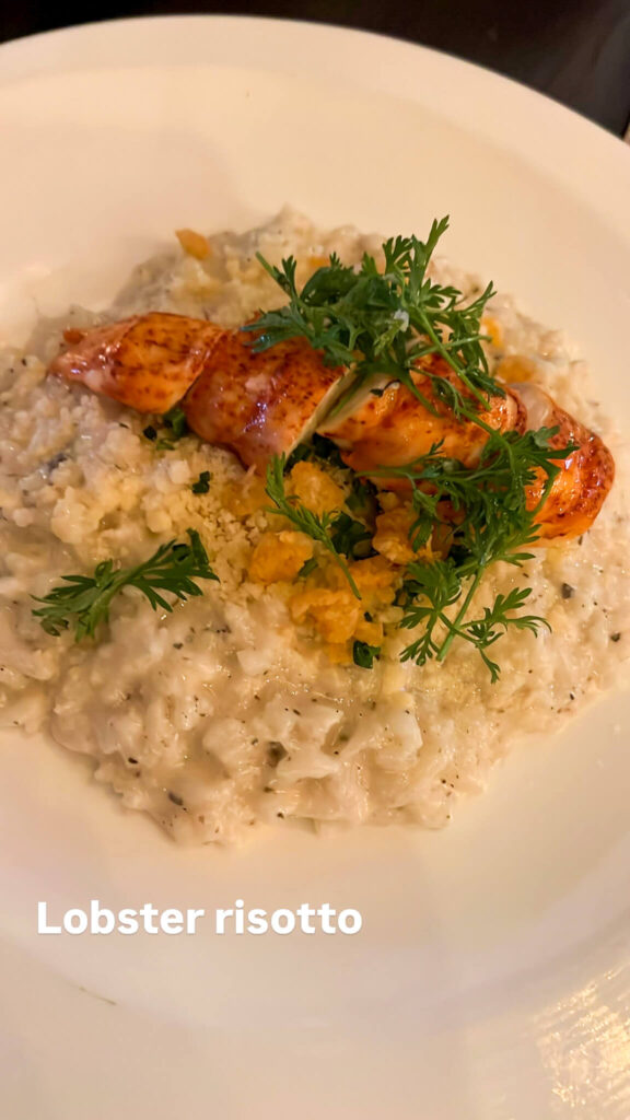 A plate of lobster risotto topped with a lobster tail and garnished with fresh herbs. The creamy risotto is served on a white dish, with the text Lobster risotto at the bottom left corner.