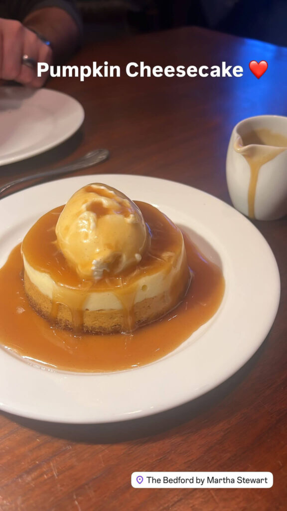 A pumpkin cheesecake topped with a scoop of ice cream and caramel sauce is served on a white plate. A small white pitcher is beside it on a wooden table. Text reads Pumpkin Cheesecake ❤️ and The Bedford by Martha Stewart.