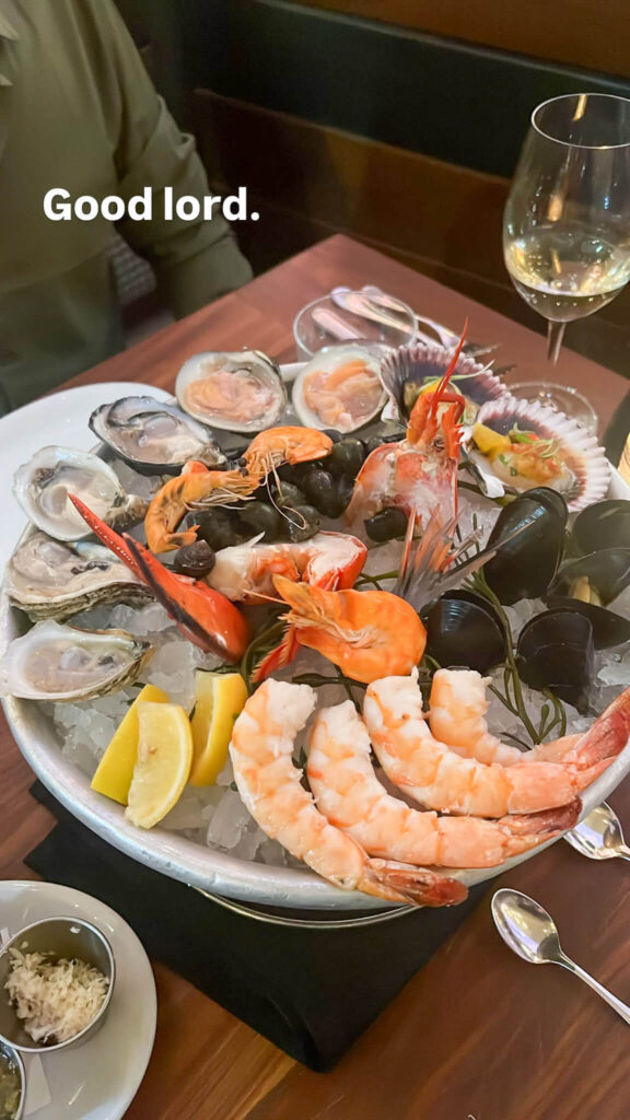 A seafood platter with oysters, shrimp, mussels, and crab legs on a bed of ice, garnished with lemon wedges. A glass of white wine and a small dish of sauce are beside the platter. The text Good lord. is displayed at the top.