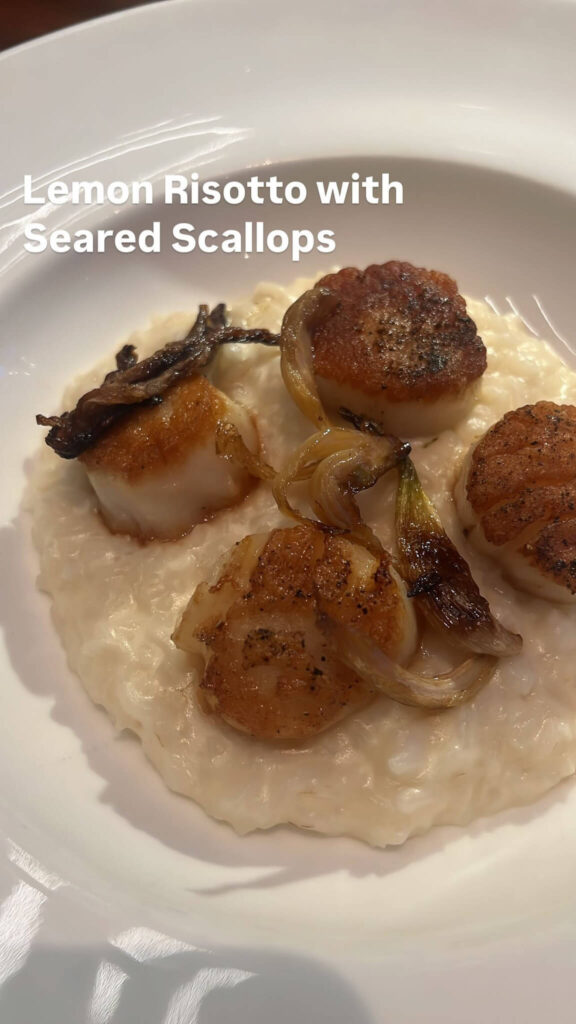 A dish of lemon risotto topped with seared scallops and garnished with caramelized onions, served on a white plate. The scallops are golden brown and arranged neatly over the creamy risotto. Text overlay reads Lemon Risotto with Seared Scallops.