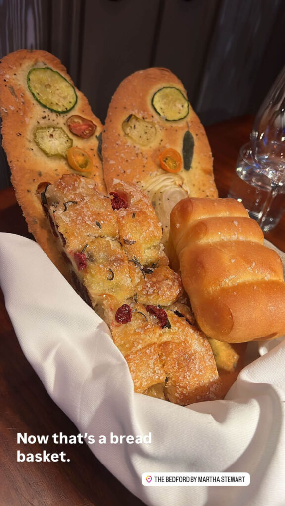 A white basket holds a variety of breads, including two long, oval-shaped loaves with sliced vegetables on top, a rectangular piece with cranberries, and three small rolls. Text reads, Now thats a bread basket. Theres a label: The Bedford by Martha Stewart.