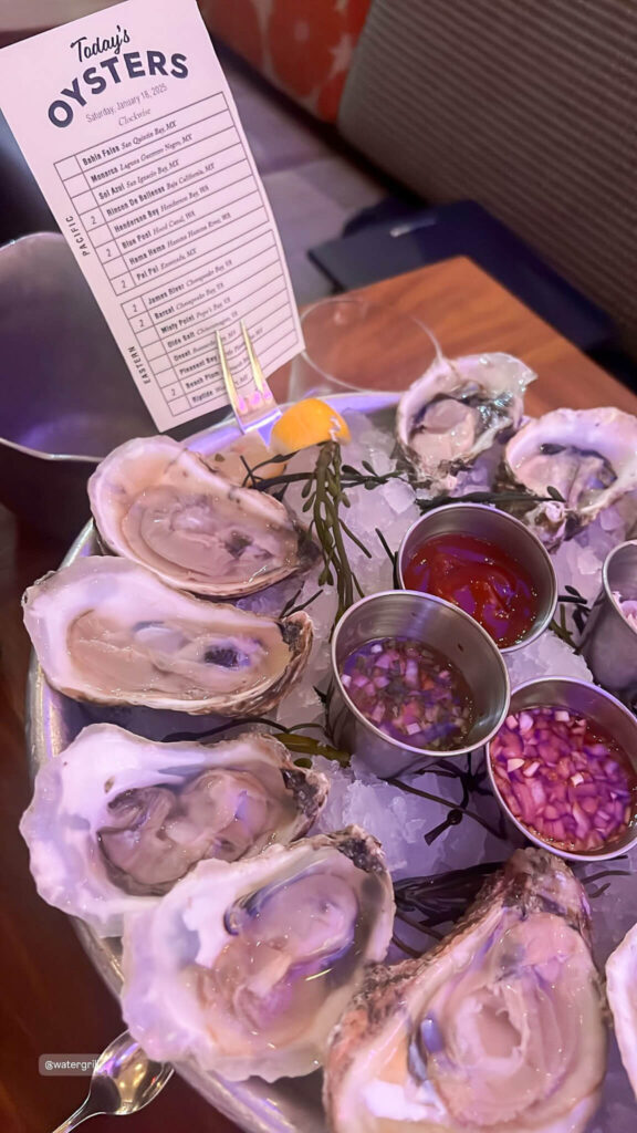 A plate of fresh oysters is arranged on a bed of ice, accompanied by condiments like lemon wedges, red sauce, and chopped onions. A menu card listing the days oyster selection is placed in the background.