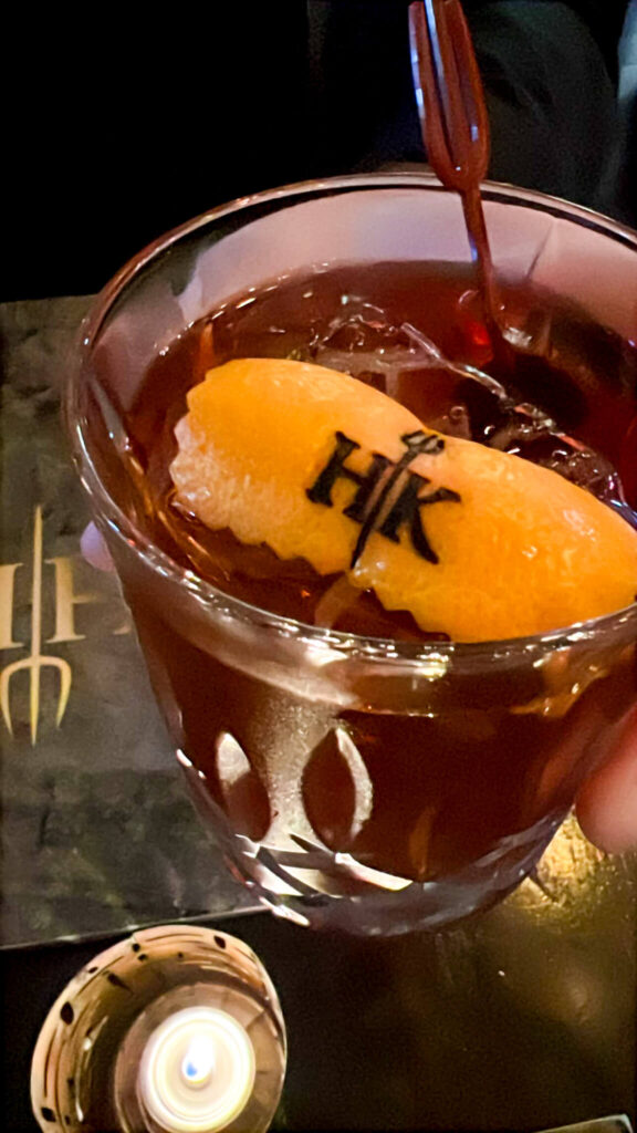 A close-up of a cocktail in a glass, garnished with an orange peel featuring a stamped HK logo. The drink is on a table with a menu and candle in the background, creating a cozy ambiance.